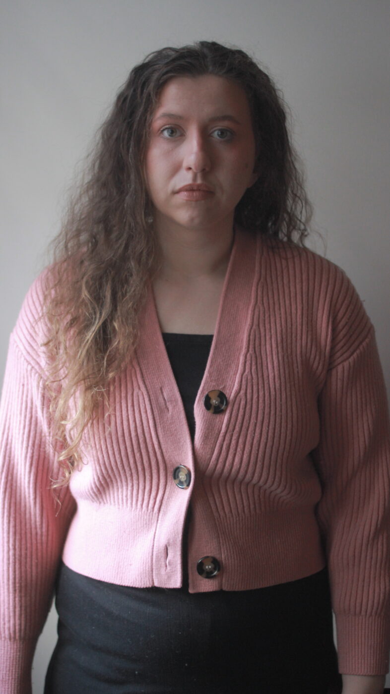 A white fem person with curly brown hair and blue eyes looks straight ahead. They are wearing a pink sweater and black dress. They stand in front of a white wall indoors against the natural light.
