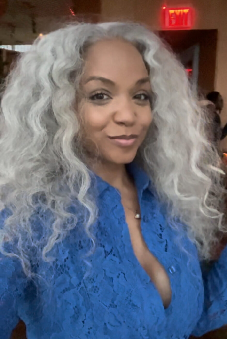 Sinnamon Love is a Black woman with a medium brown complexion, long wavy grey hair, and is wearing a blue lace dress with buttons and an open collar.