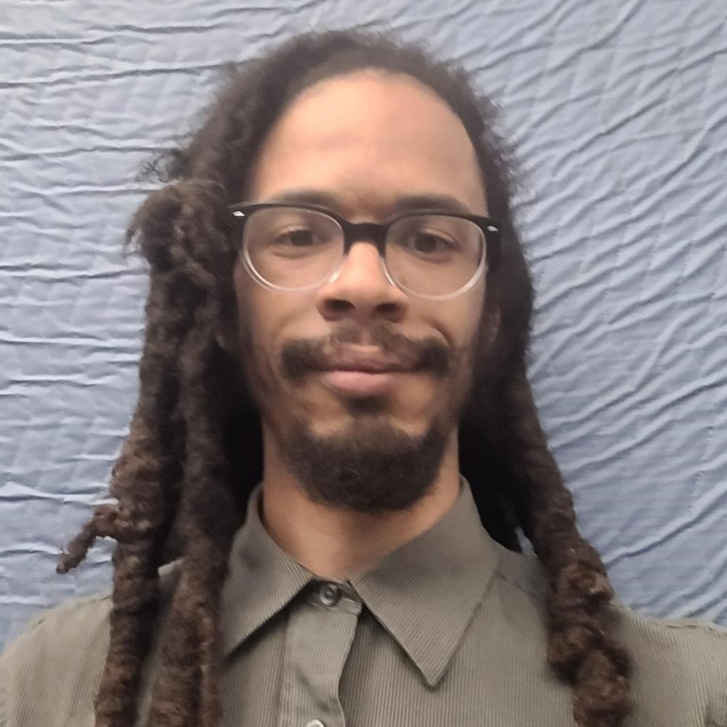 Image is of a medium-skinned black man with a beard and mustache. He has long dreadlocks and is wearing glasses.