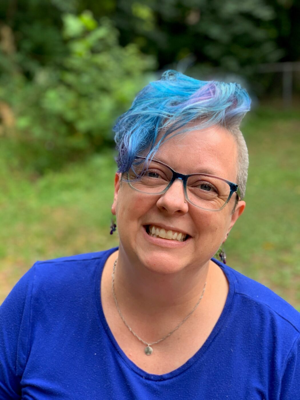 Chris Ash, a light-skinned white queer person smiles in their backyard. They have a blue shirt, short blue and purple hair, and blue glasses.
