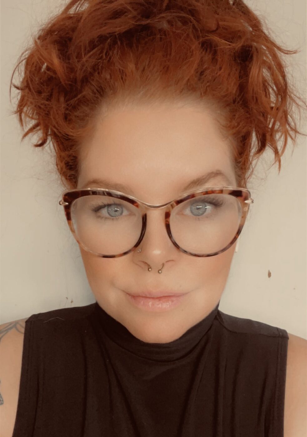Rebekah has fair skin with tattoos on her arm, and is wearing a black turtleneck, with red tortoiseshell glasses and her hair is reddish-brown and pulled up in a bun.