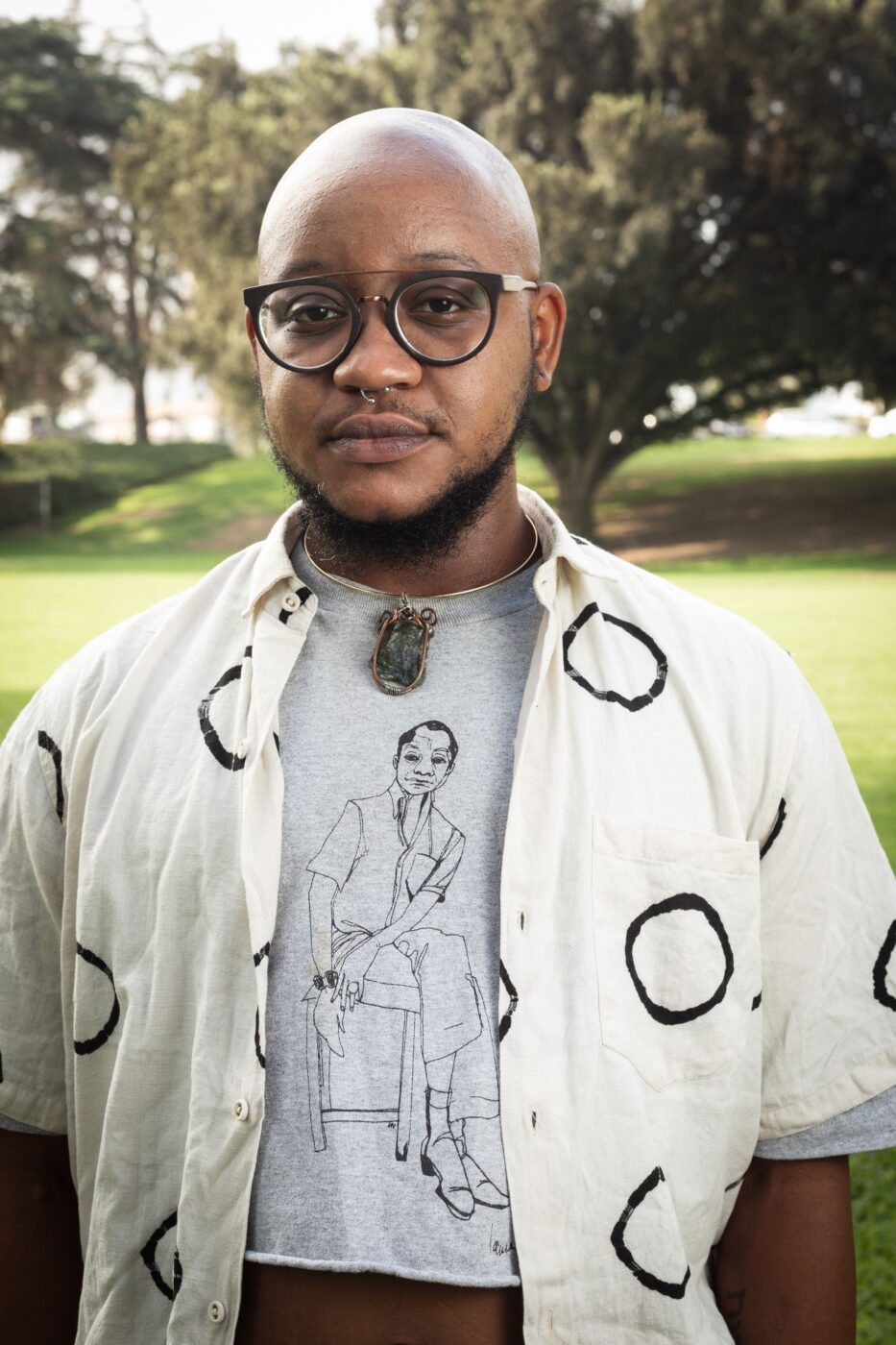 Jaden, a brown-skinned black trans man with a bald head, round glasses, a septum ring, wearing a light gray cropped shirt with an illustration of James Baldwin, a light beige unbuttoned shirt with Black circles on it, and a Labradorite crystal choker necklace.