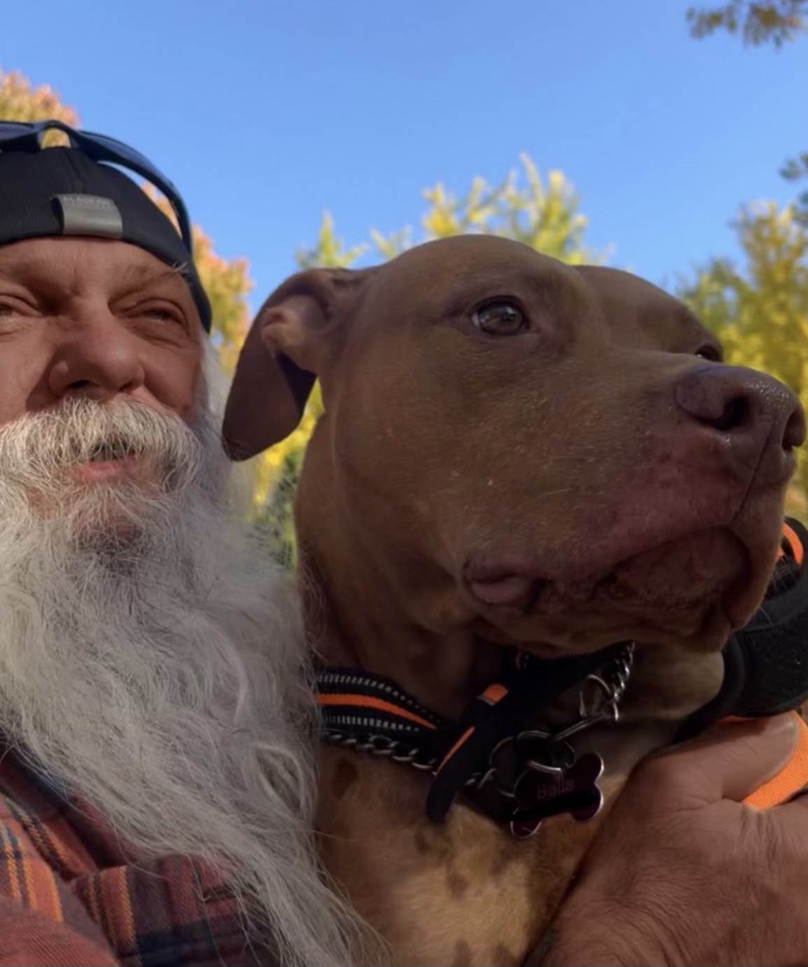 A brown-skinned man with a long white beard snuggles an adorable dog. The man is wearing a cap.
