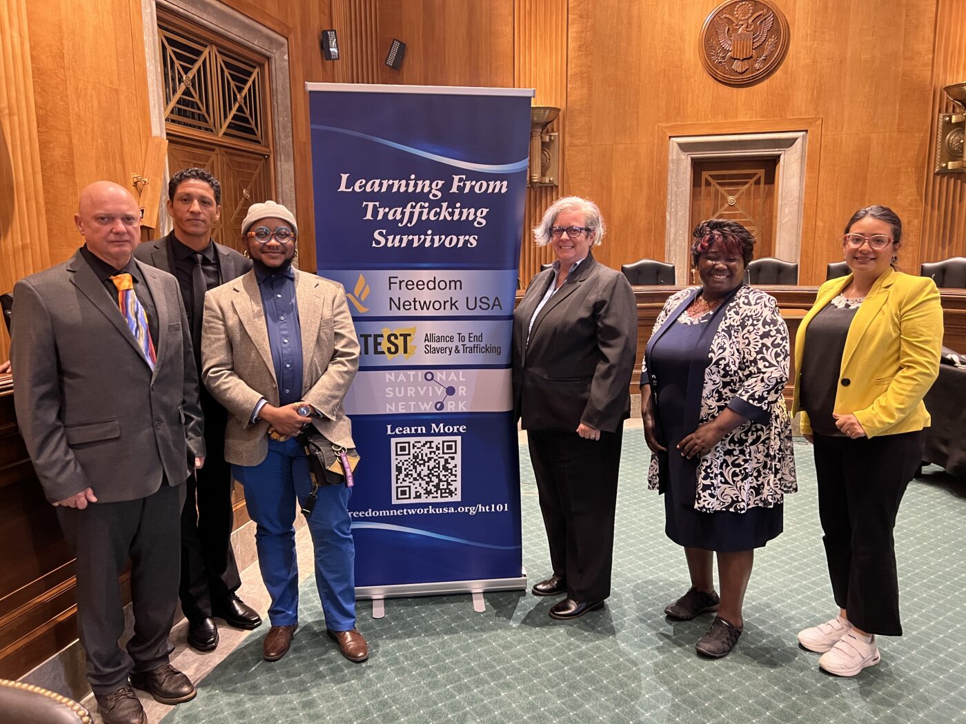 Six stunning experts with lived experience stand next to a banner in the Senate Foreign Relations Committee Hearing Room. The banner reads Learning from Lived Experience